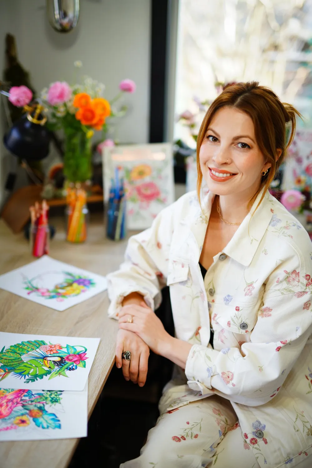 Rachel Reinert seated at her studio table with fresh flowers and colored pages